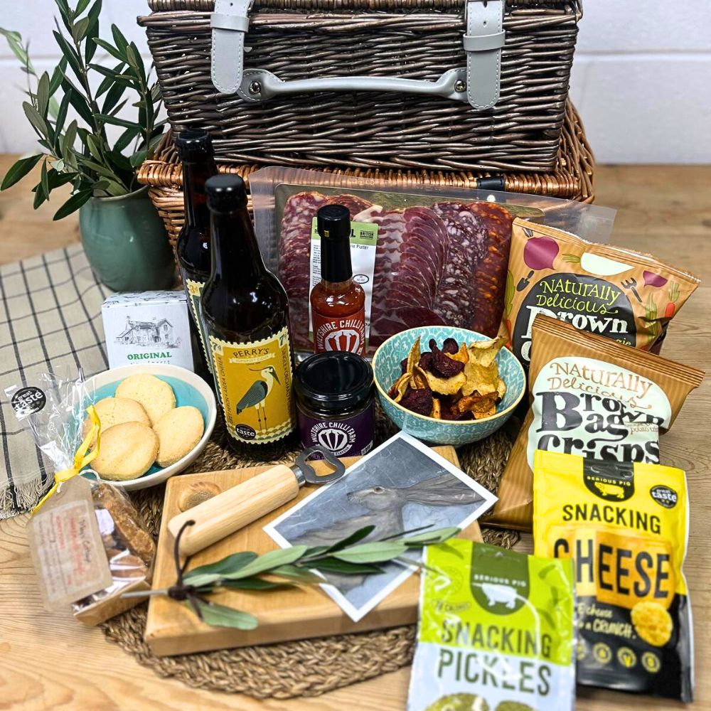 Picnic basket with food items including cheese, pickles, and meats on a wooden table.