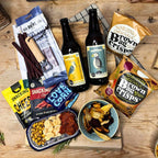 A display featuring a match pack next to a hamper overflowing with snacks, paired with bottles of cider. Several of the snacks are taken out and neatly presented on plates in front of the hamper.