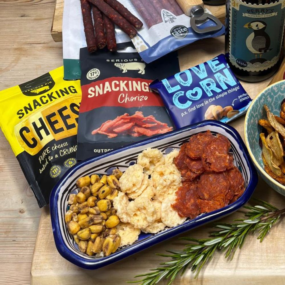 Assorted snacks including cheese, chorizo, and corn on a wooden surface with packaging in the background.
