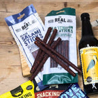 Assorted food items including salami sticks, venison sticks, and a bottle of Perry's cider on a wooden surface.